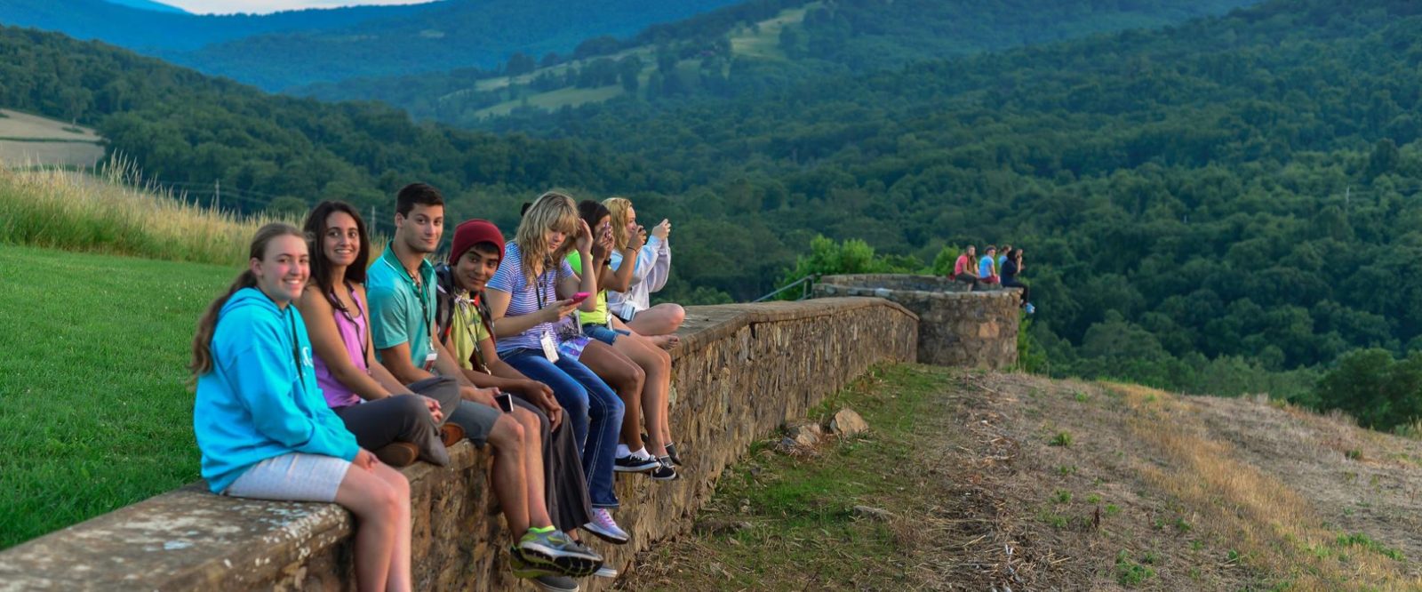 Students sit along a stone wall looking out over a mountain view