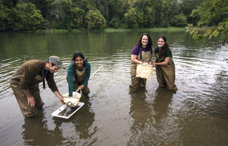 Students in waders smile at camera standing in river collecting samples